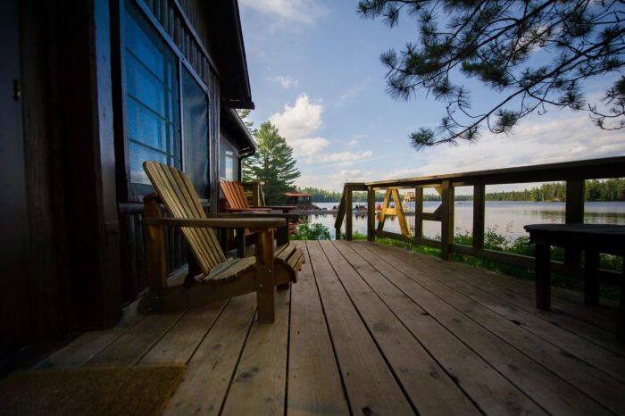 Two Muskoka chairs on a wooden deck facing a lake. In the background there’s a pier