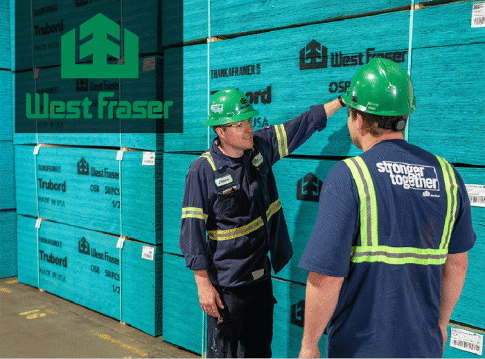 Two West Fraser employees wearing safety gear talk in front of stacked blue Trubord OSB panels inside a warehouse. One employee gestures toward the product, while the other wears a shirt that says ‘stronger together.