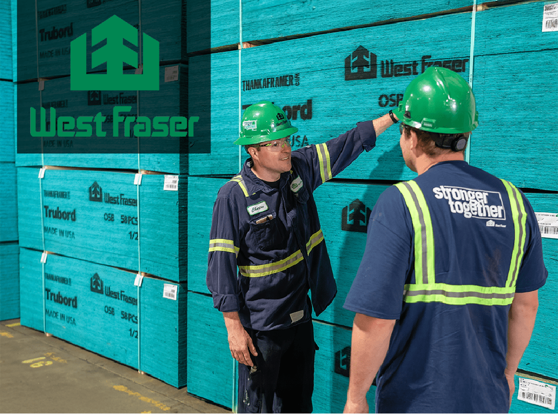 Two West Fraser employees wearing safety gear talk in front of stacked blue Trubord OSB panels inside a warehouse. One employee gestures toward the product, while the other wears a shirt that says ‘stronger together.