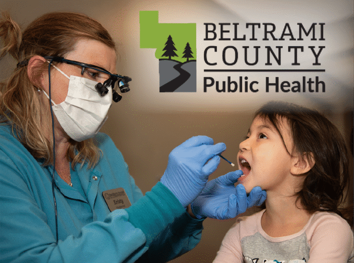 A dental worker wearing a mask examines a young child’s mouth during a dental checkup. The Beltrami County Public Health logo appears in the upper right corner