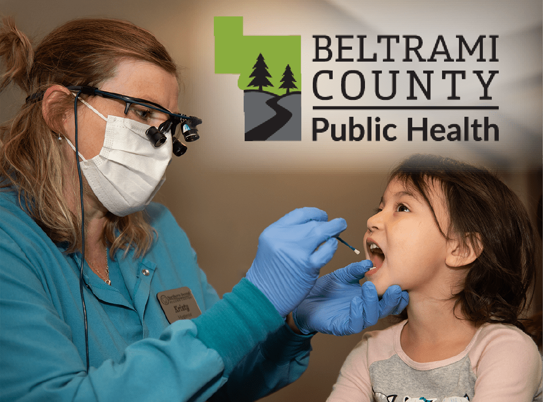 A dental worker wearing a mask examines a young child’s mouth during a dental checkup. The Beltrami County Public Health logo appears in the upper right corner