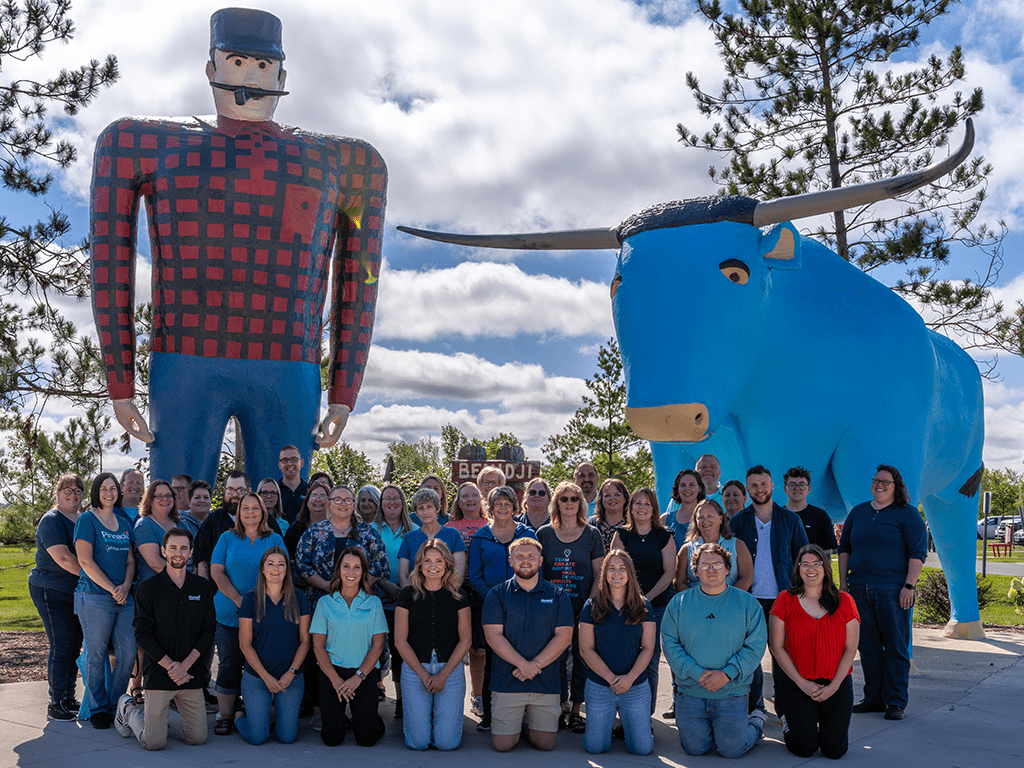 A large group of people from Pinnacle Marketing Group pose together outdoors in front of the iconic Paul Bunyan and Babe the Blue Ox statues in Bemidji, Minnesota. The team stands and kneels in rows, smiling on a bright day with scattered clouds and tall pine trees in the background.